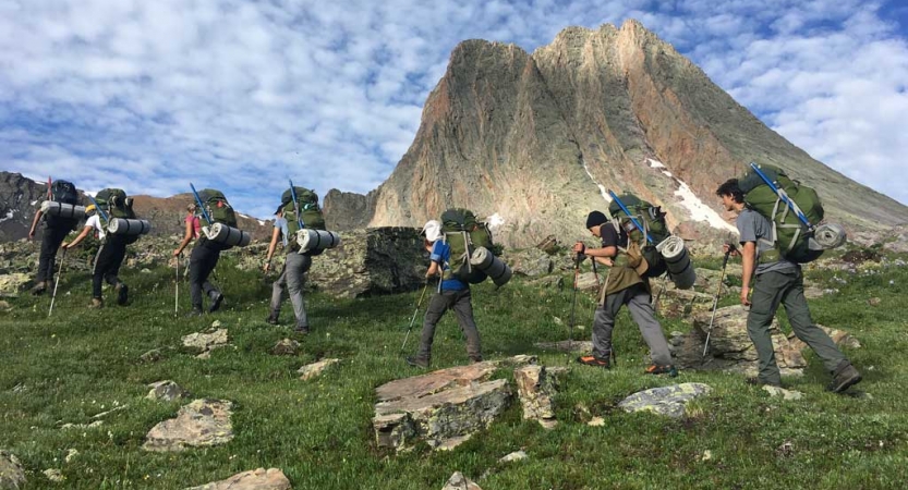 A group of backpackers cross green and rocky terrain with a mountain in the background. 